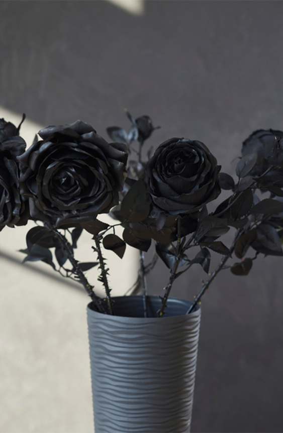 Elegant bouquet of black long-stem roses in textured, gray vase against a gray wall background. Light shines in from the left through a window not seen in image.