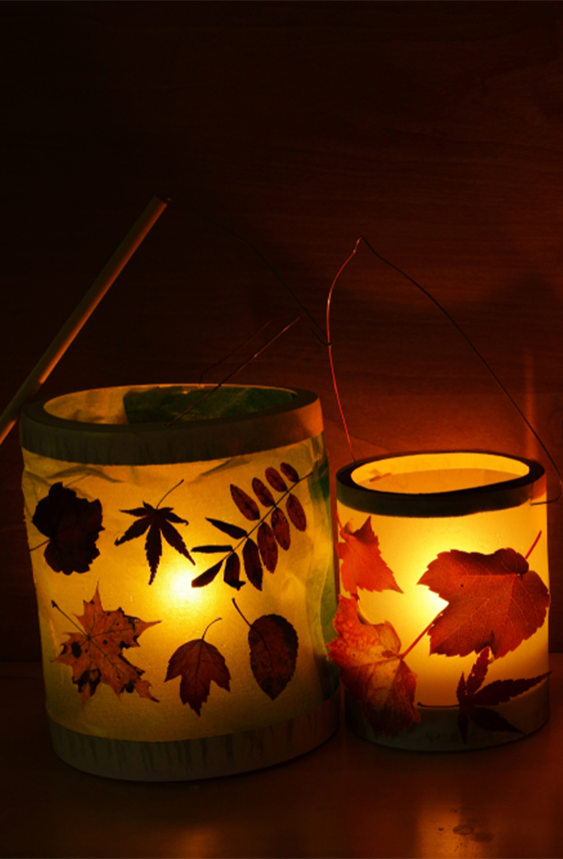 Homemade lanterns wrapped in tissue paper with various pressed leaves glued on. The jars sit on a dark cherry wood shelf and are illuminated by tea candles inside.