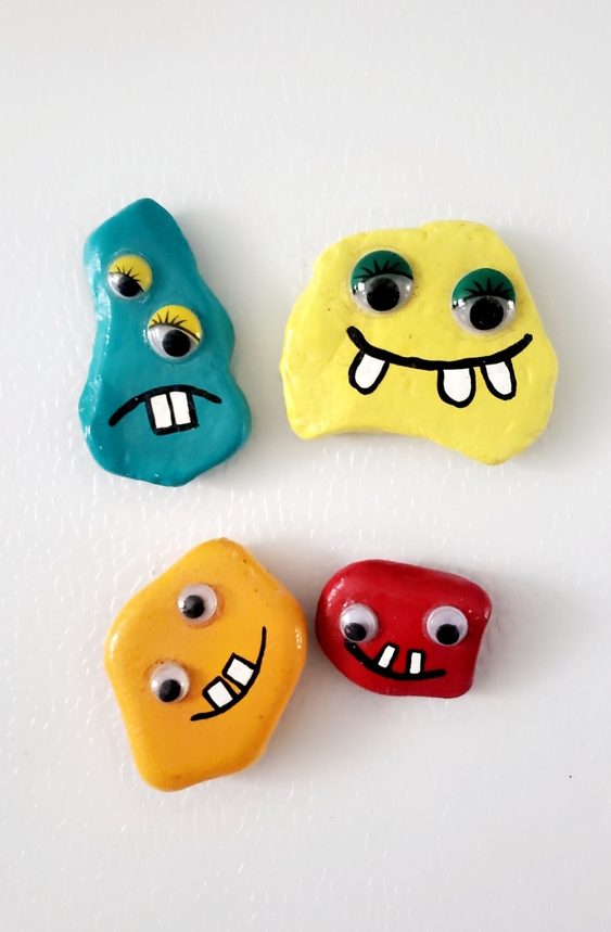 Four misshapen rocks painted in varying bright colors, each with different style and size googly eyes glued onto them, and silly mouths with buck teeth painted on. Top view against an off-white table background.